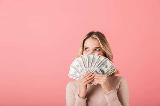 A smiling woman holding hundred dollar bills as a fan in her hand in front of a pink background