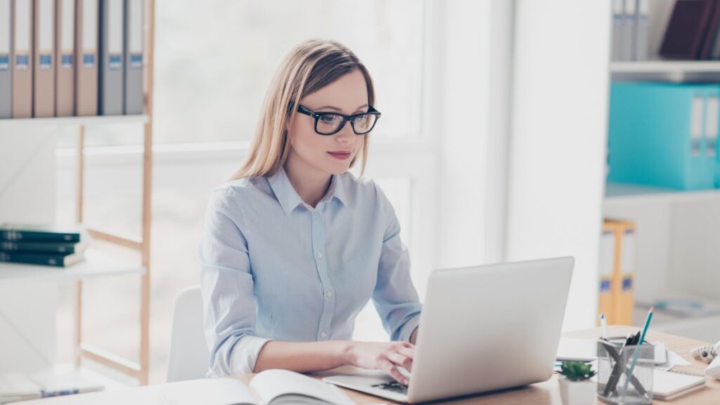 A woman in glasses typing on a desktop while dressed professionally in a light blue collared shirt