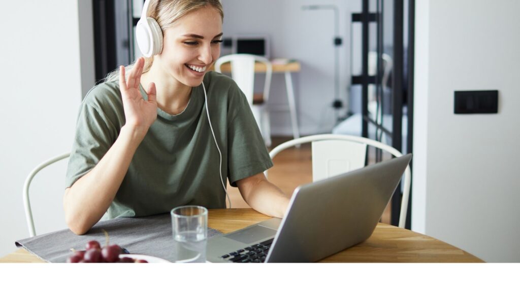 a blonde woman wearing headphones teaching English online at her desk smiling and waving at her student on her laptop 