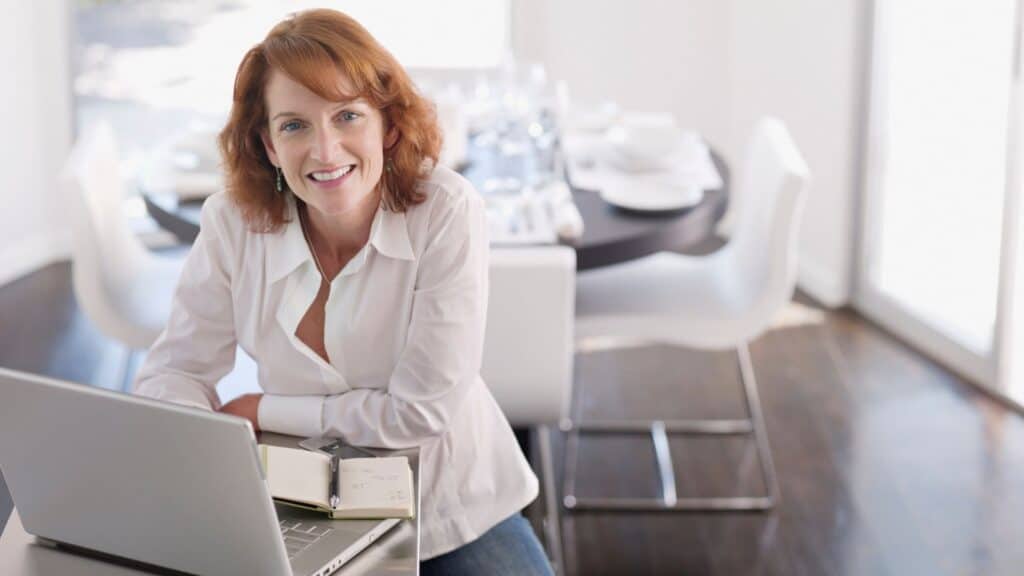 a woman with red hair in a white collared shirt smiling while sitting at her desk in front of her laptop in a room full of white furniture 