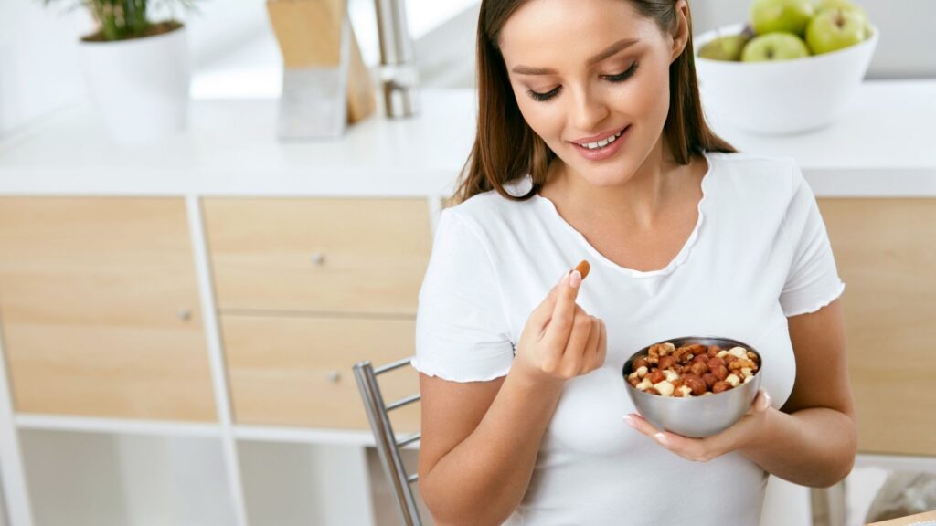 a woman in a white shirt smiles and looks down at nuts in her hand while holding a bowl of nuts in her other hand