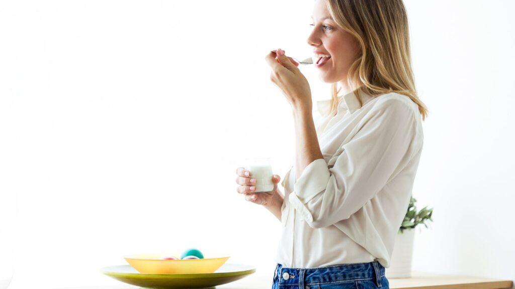the profile of a blonde woman in a white shirt and jeans eating yoghurt in a white room 