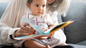 An adult with a baby on her lap helping the baby learn English as a second language by reading a book with her while sitting on a couch