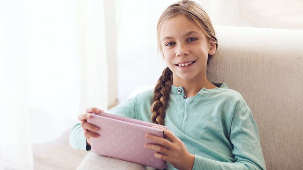 a smiling young girl with a long braid holding her tablet with a pink case on her lap 