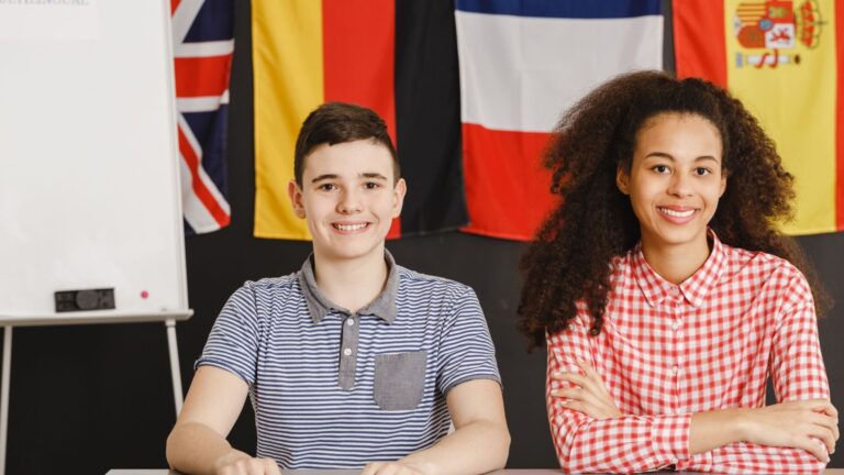 a photo of two young English students sitting in front of various country flags with their arms crossed and smiling