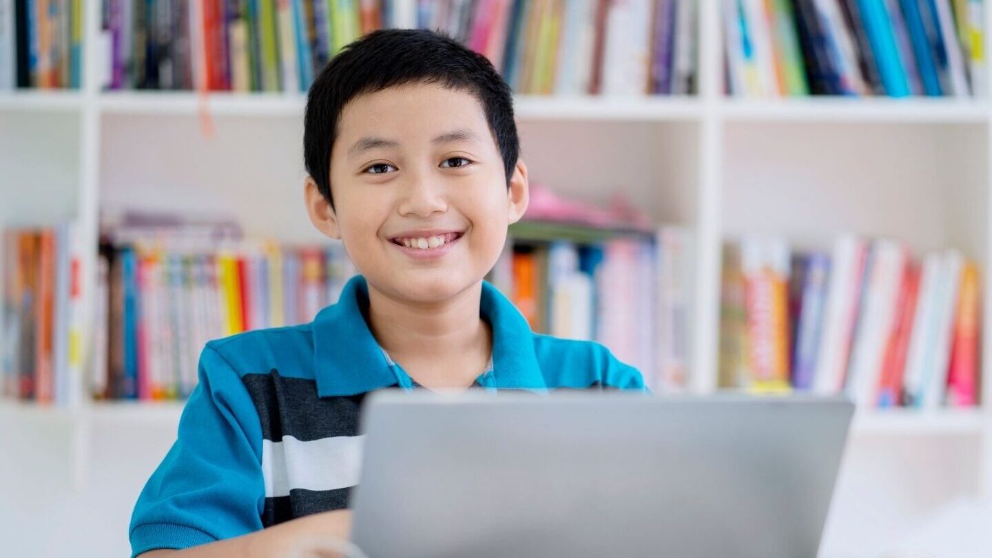 an asian boy in a blue shirt smiling in front of his laptop 