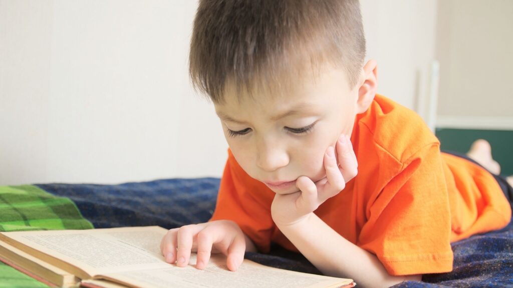a young kindergarten age boy laying on his stomach reading a book while resting his head in his hands 