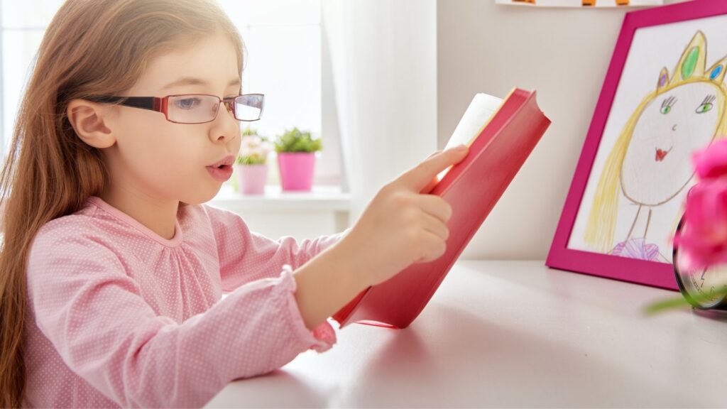 a young girl  in glasses reading a book and mouthing the sounds as she reads