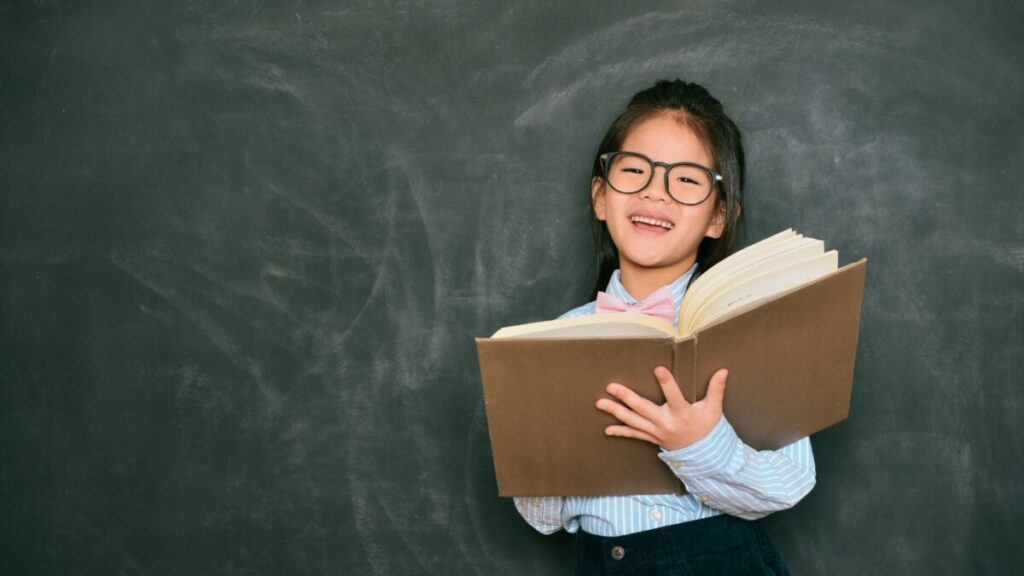 a young asian girl holding a book open in front of a blackboard and smiling 