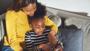a young blank mom leans over her black daughter and points at a screen helping her daughter learn to read 