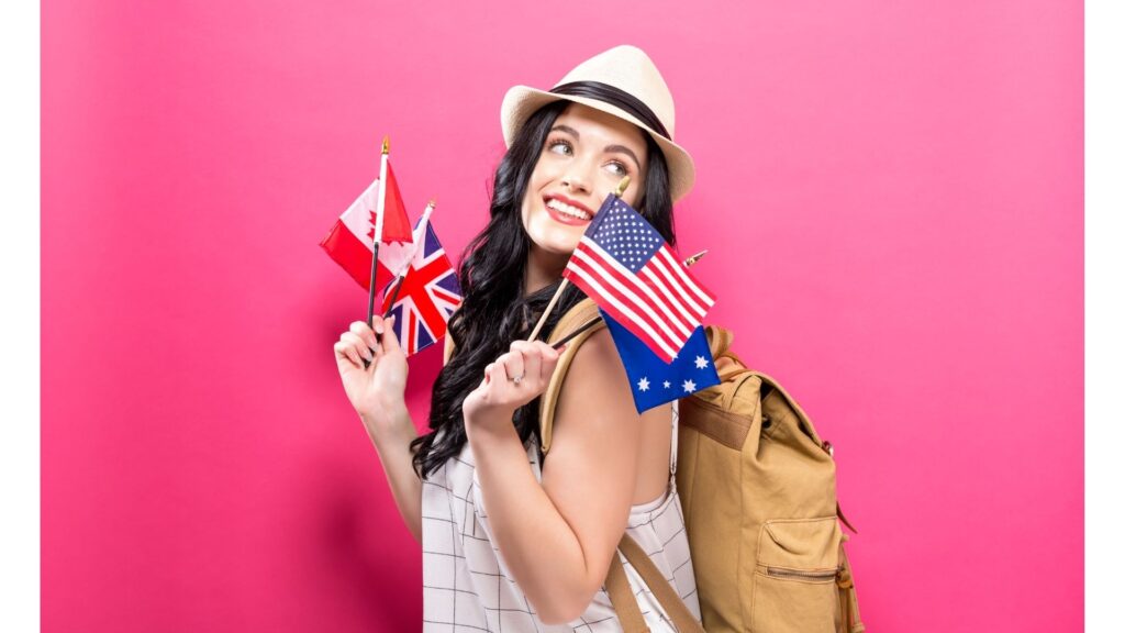 A smiling woman in a hat and wearing a backpack holds many foreign flags in her hands while standing in front of a pink background 