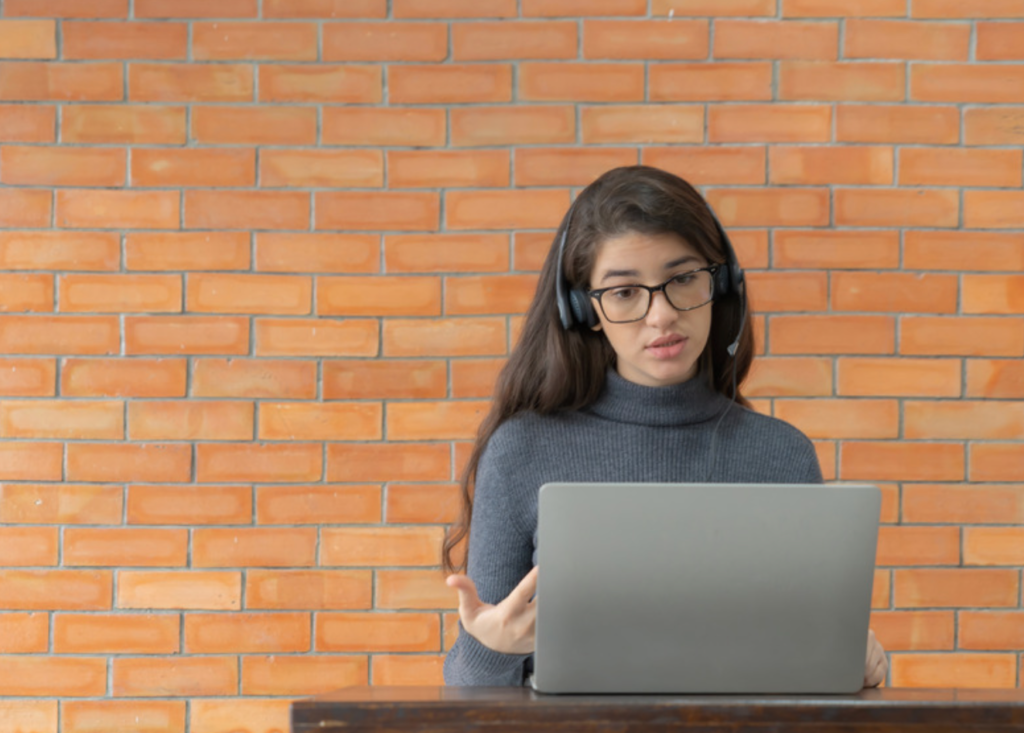 a brunette woman with long hair sitting in front of a brick wall at a desk and talking to someone on a laptop while wearing headphones 