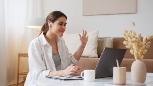 A woman smiling and waving at her laptop while sitting in a beige room