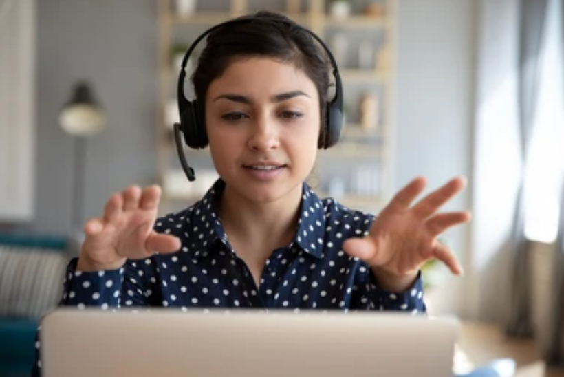 a photo of a woman with dark brown hair wearing headphones using her hands to gesticulate while talking to someone on her laptop